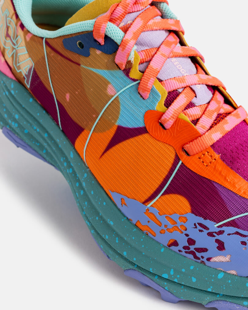 Close-up of a colorful running shoe with a multicolored upper, speckled teal sole, orange accents, and coral pink laces. The brand name "HOKA" is visible on the side