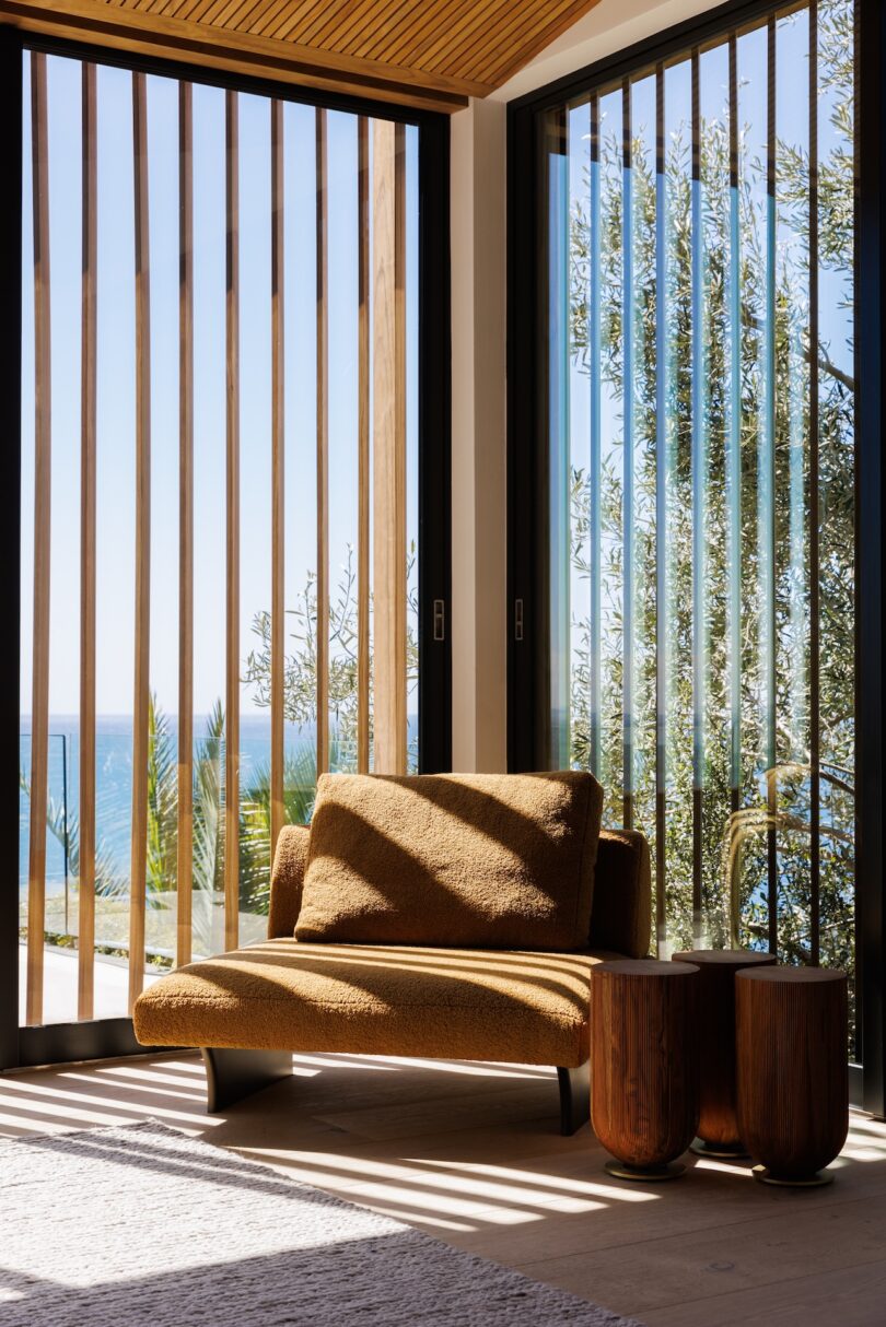 A brown armchair and a small wooden side table are positioned by floor-to-ceiling windows with vertical wooden slats, overlooking trees and distant ocean views.