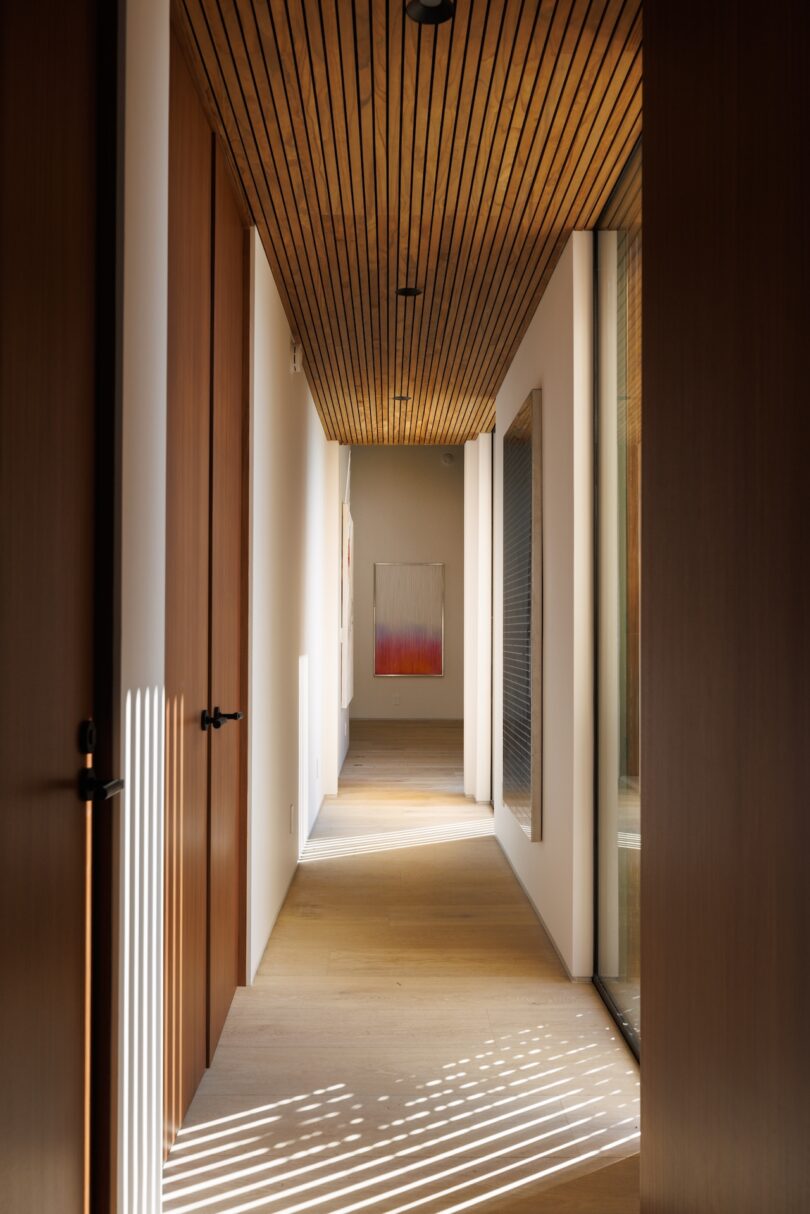 A modern hallway with wooden ceiling and doors, large windows on one side, and a framed abstract artwork at the far end, illuminated by natural light.