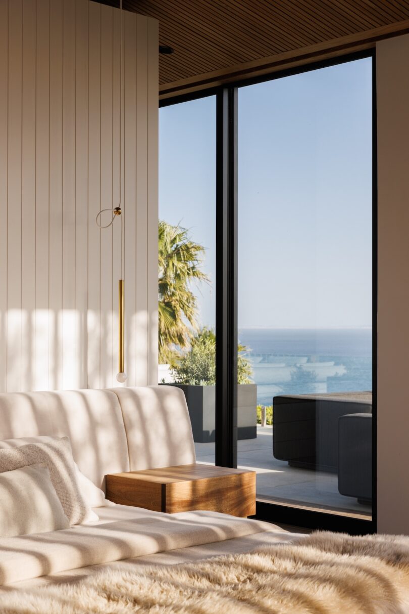 Modern bedroom with a bed, wooden side table, and large window offering a view of the ocean, outdoor seating, and palm trees in natural daylight.