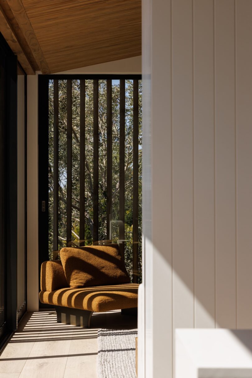 A modern lounge chair with mustard upholstery sits in a sunlit corner near vertical slatted windows, casting striped shadows on the floor.