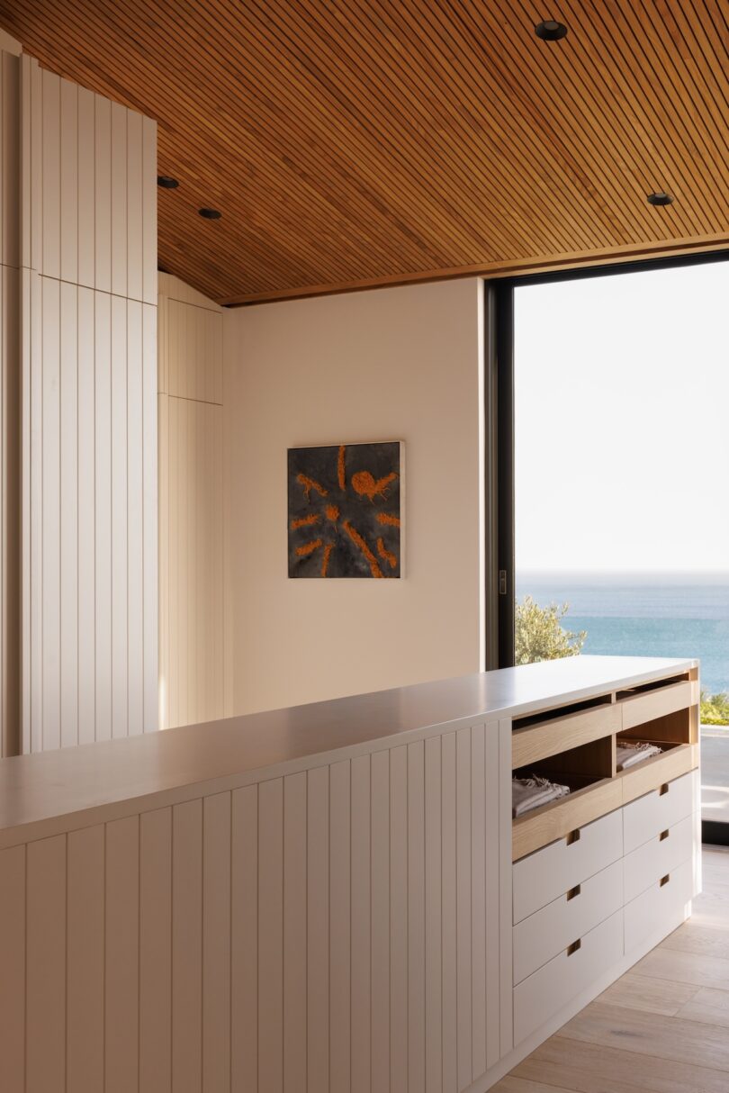Modern kitchen with white paneled cabinets, open shelving, wood slat ceiling, wall art, and large window with an ocean view.