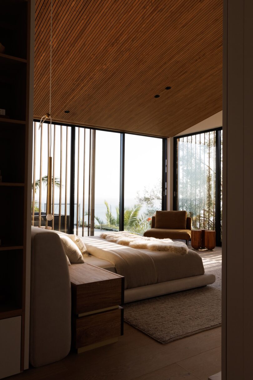 A modern bedroom with a wooden ceiling, large glass windows, a neatly made bed, a beige armchair, and natural sunlight streaming in.