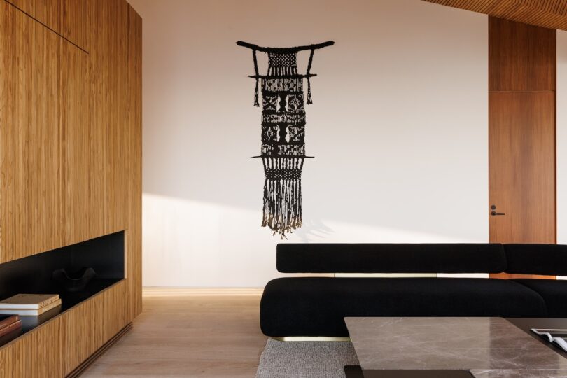 A modern living room with wooden paneling, a black sofa, a marble coffee table, and a black woven wall hanging displayed on a white wall.