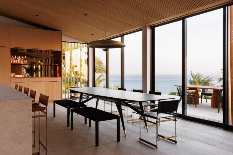 Modern dining room with a black table, minimalist chairs, large windows overlooking the ocean, and wood-paneled ceiling.