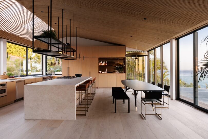Modern kitchen and dining area with wood panel ceiling, large island, minimalist furniture, floor-to-ceiling windows, and views of greenery and water outside.
