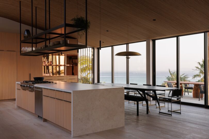 Modern kitchen and dining area with light wood finishes, marble island, open shelving, large windows, and an ocean view.