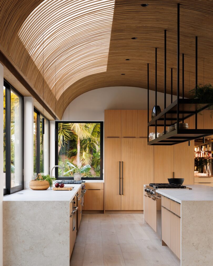 Modern kitchen with light wood cabinetry, a stone island, large windows showing greenery, and a curved wood slat ceiling.