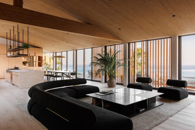 Modern open-plan living room with black sofas, a large coffee table, indoor plants, and floor-to-ceiling windows offering an ocean view. Wooden ceiling and accents throughout.