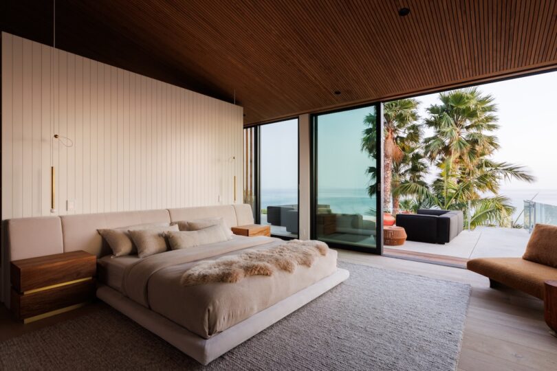Modern bedroom with a large bed, neutral decor, wood-paneled ceiling, and floor-to-ceiling windows opening to a patio with palm trees and ocean view.