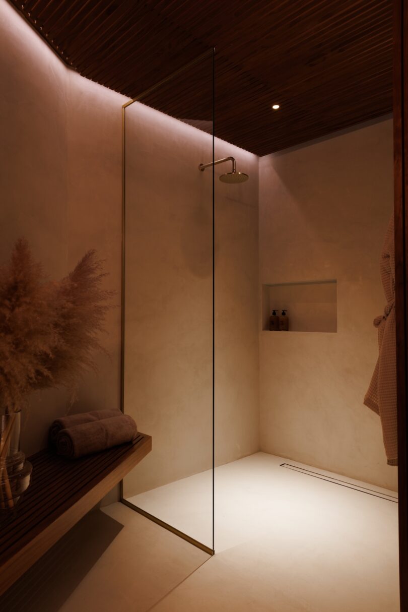 Minimalist walk-in shower with glass partition, wall-mounted showerhead, recessed shelf with toiletries, wooden bench, rolled towels, and pampas grass decoration, lit by warm ambient lighting.
