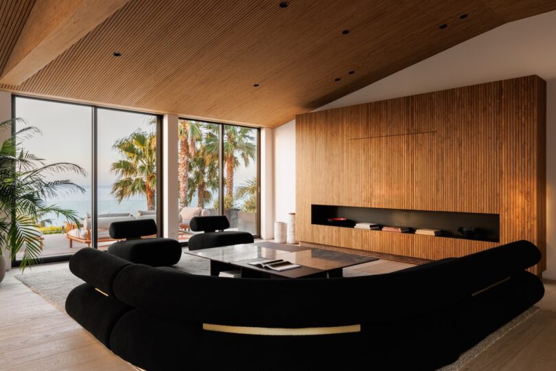 Modern living room with black curved sofa, wood-paneled walls and ceiling, large windows, and a view of palm trees and the ocean outside.