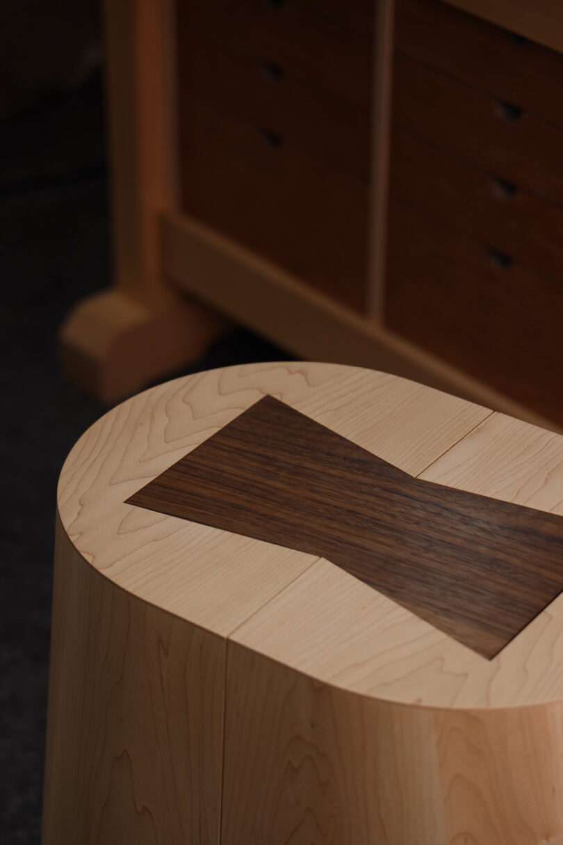A wooden stool made of light-colored wood features a dark wood butterfly joint inlay on the seat; a wooden cabinet is in the background
