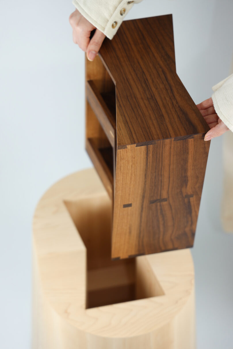 A person places a small, rectangular wooden box with shelves into a matching slot on a light-colored wooden base