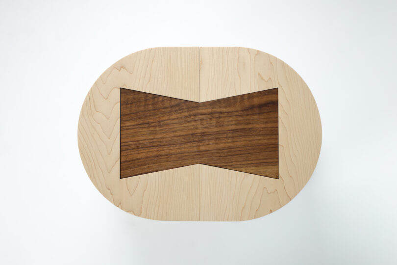 Oval wooden stool with a contrasting dark wood bowtie-shaped inlay in the center, viewed from above against a plain white background