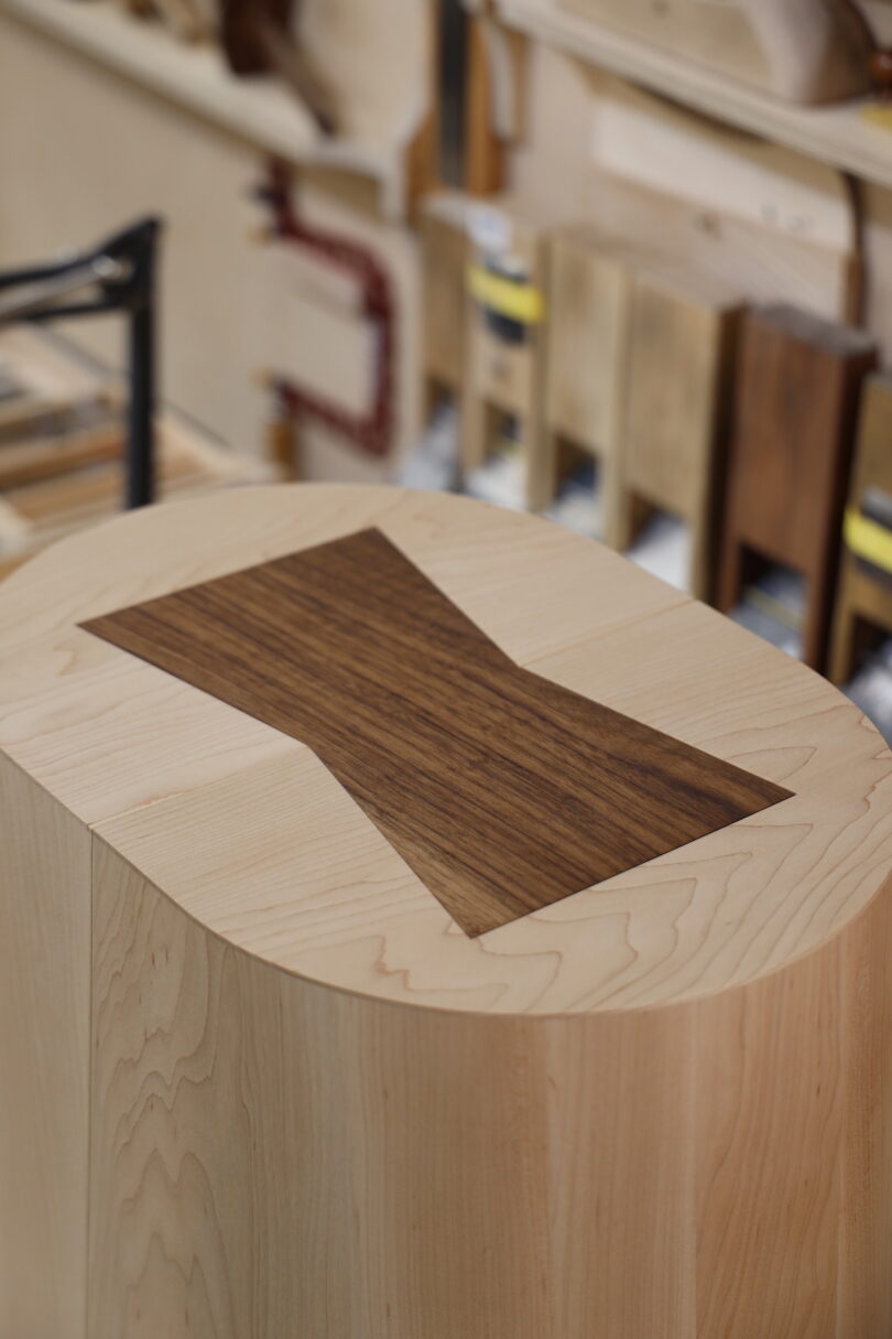 A close-up of a wooden furniture piece featuring a contrasting butterfly joint inlay on a light wood surface, with woodworking tools and materials in the background