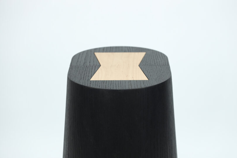 Black, asymmetrical wooden stool with a flat, light wood inlay on the top, set against a plain white background