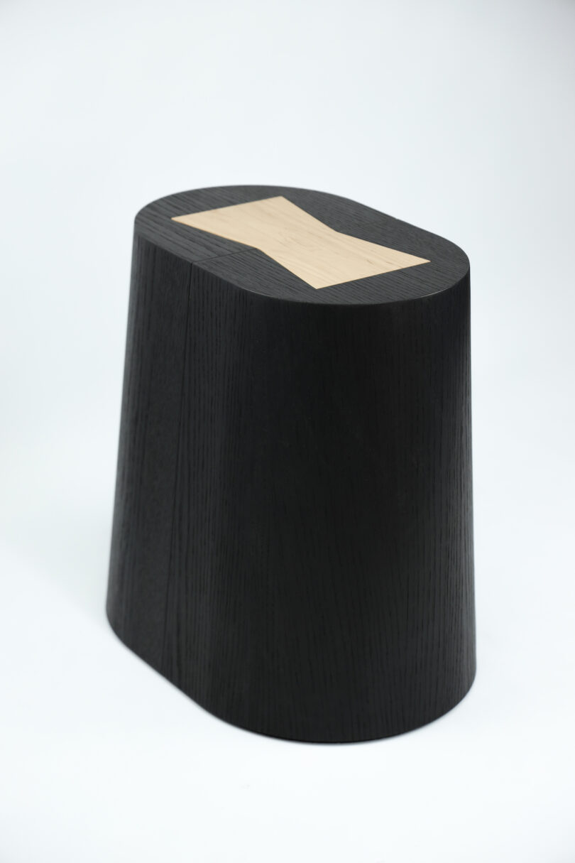 Black, asymmetrical wooden stool with a flat, light wood inlay on the top, set against a plain white background