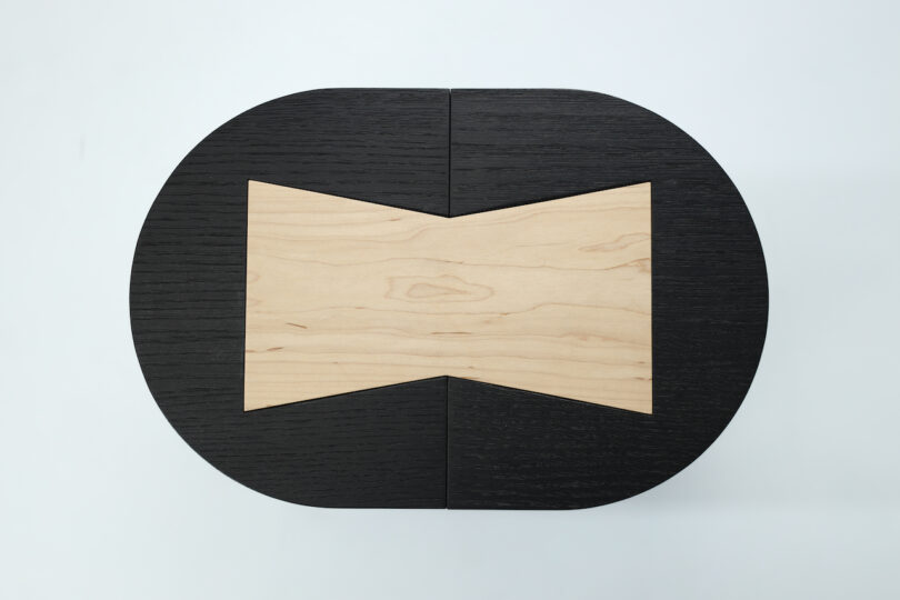 Black, asymmetrical wooden stool with a flat, light wood inlay on the top, set against a plain white background