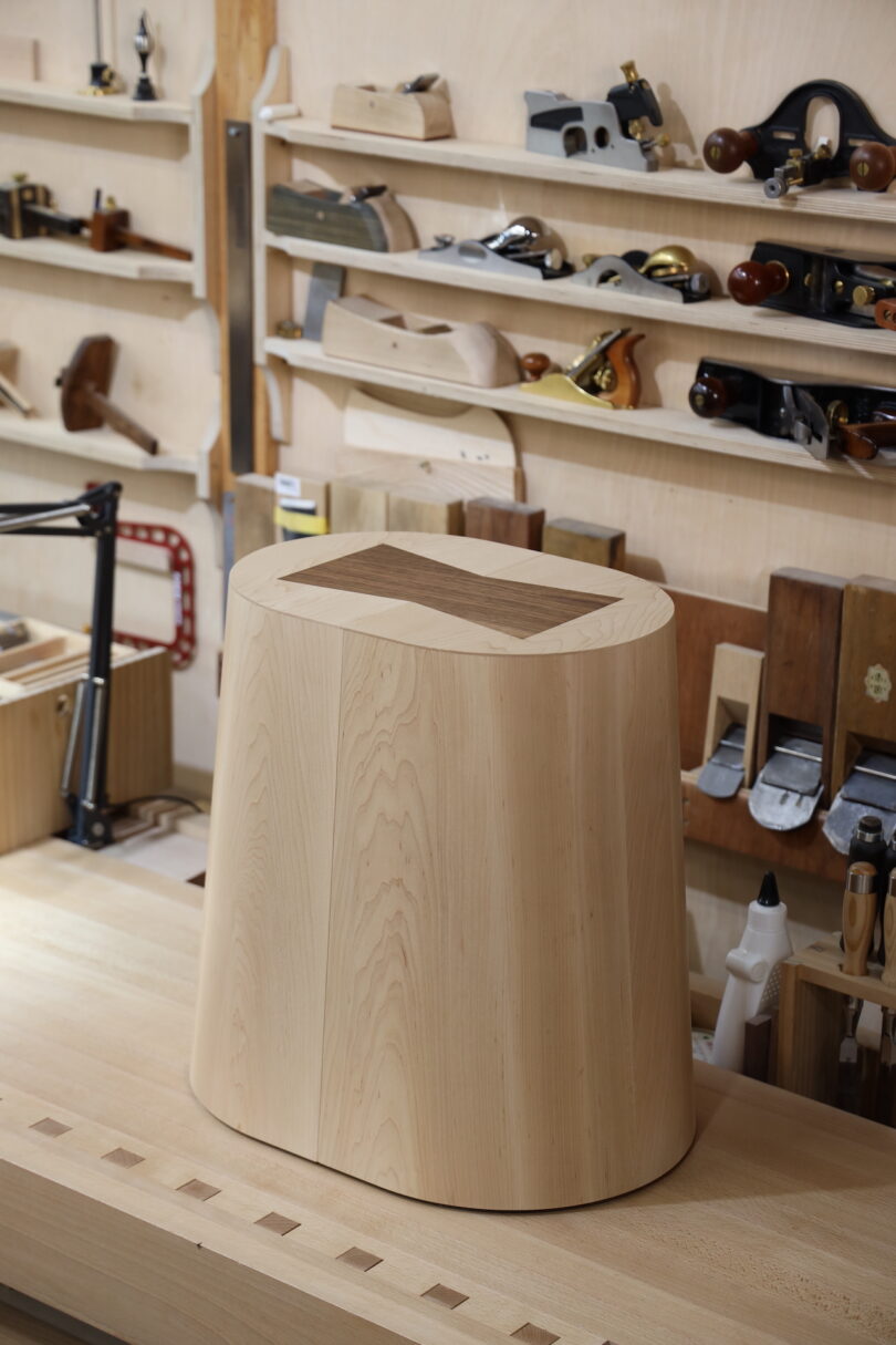 A wooden container with rounded edges sits on a woodworking bench, surrounded by various hand tools and woodworking supplies on shelves in the background