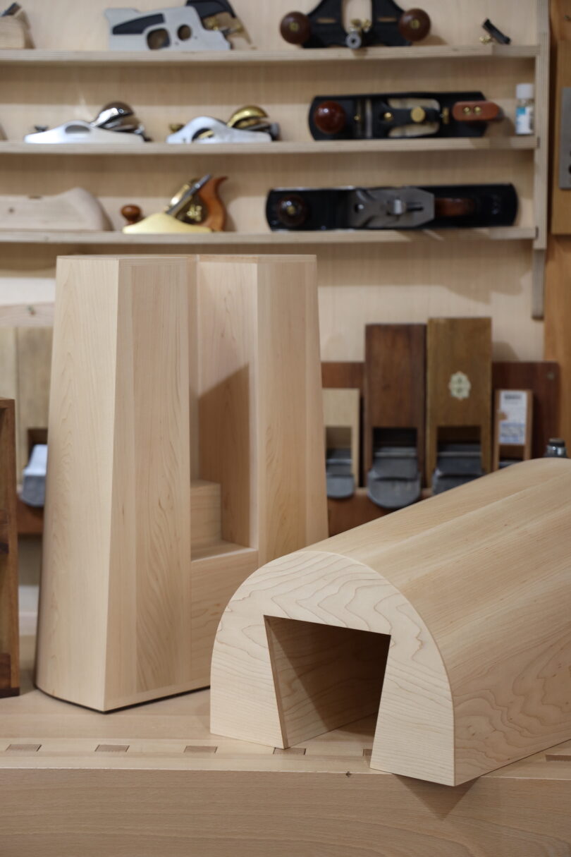 Two unfinished wooden furniture pieces sit on a workbench in a woodworking shop, with hand tools arranged on shelves in the background