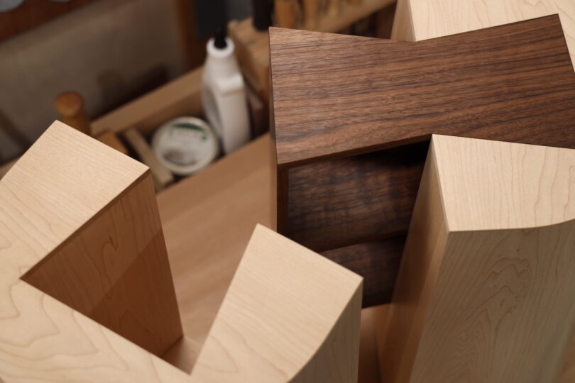 Close-up of two wooden furniture pieces with geometric cutouts, one made of light wood and the other of dark wood, in a workshop setting with tools and supplies in the background