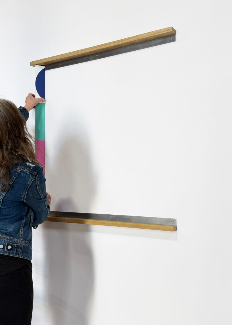A person installs or adjusts a vertical strip with colored sections and a semicircle shape between two parallel brass rails mounted on a white wall
