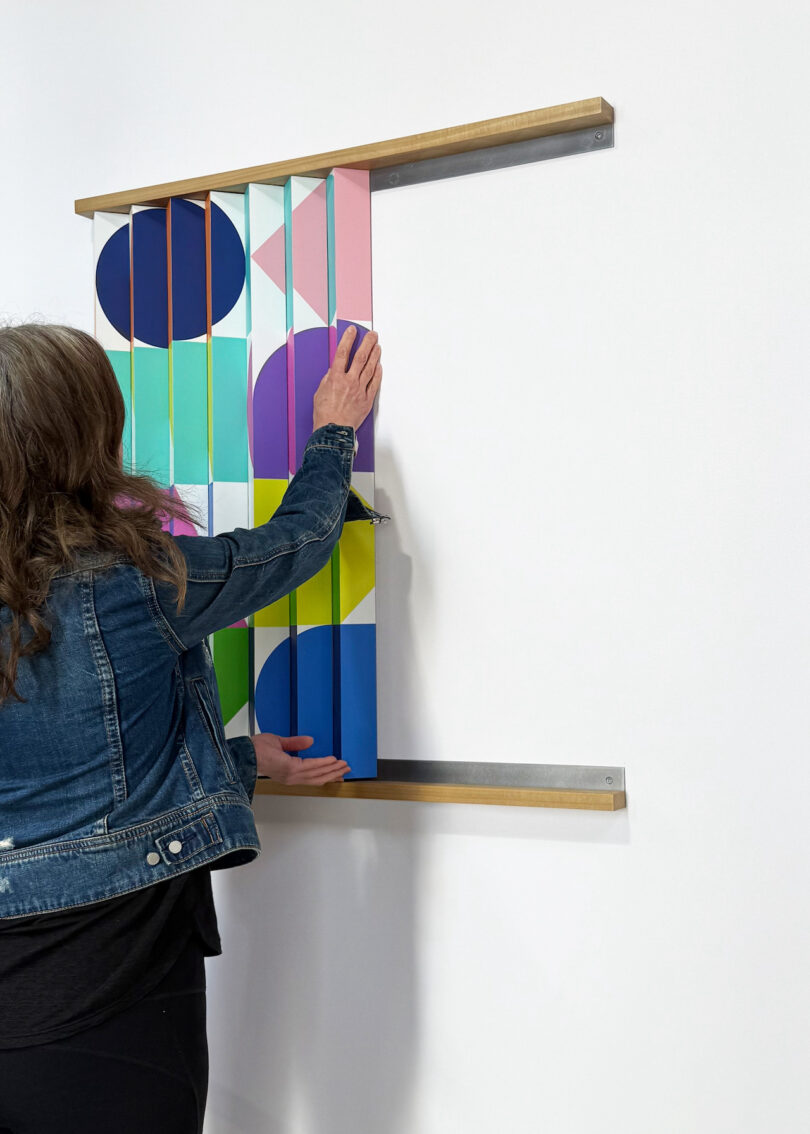 A person wearing a denim jacket adjusts a wall-mounted interactive artwork with colorful geometric shapes and vertical slats