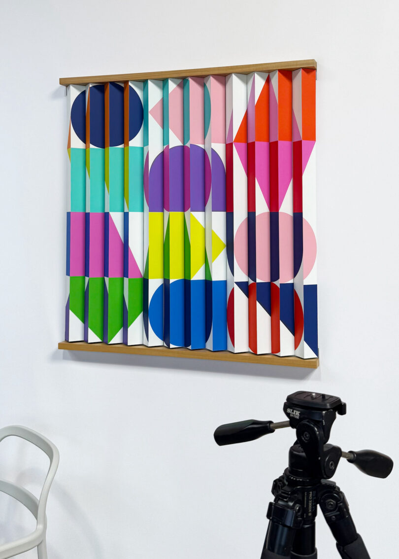 A colorful geometric artwork with vertical slats hangs on a white wall; a black tripod and part of a white chair are visible in the foreground