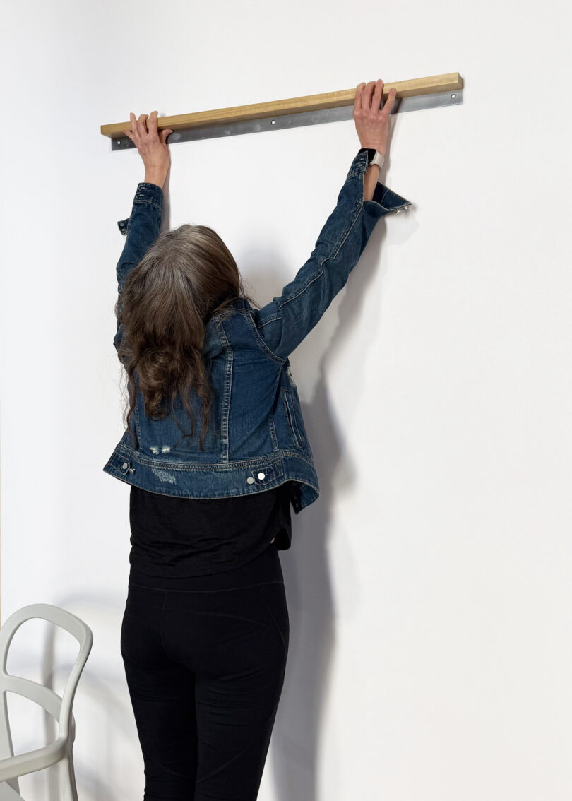 A person in a denim jacket holds onto a wooden bar mounted on a white wall, standing on the floor next to a light-colored chair
