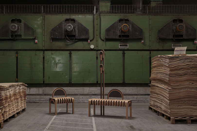 Three uniquely designed wooden chairs are arranged in an industrial setting, flanked by stacks of plywood sheets and large green industrial machines in the background