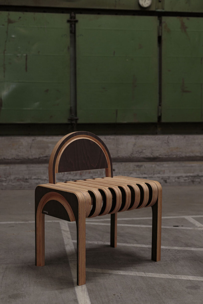 A modern chair made of layered wood with arched legs and a curved backrest, positioned in an industrial setting with a green metal wall