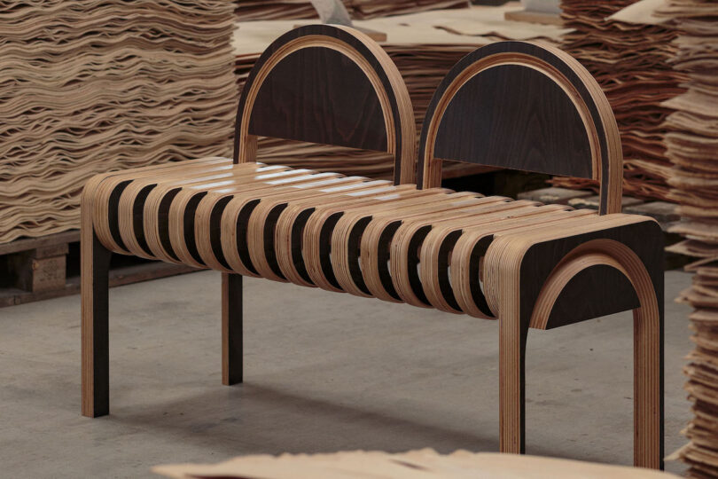 A modern wooden bench with curved, slatted seat and two semicircular backrests, surrounded by stacks of wood sheets in a workshop setting