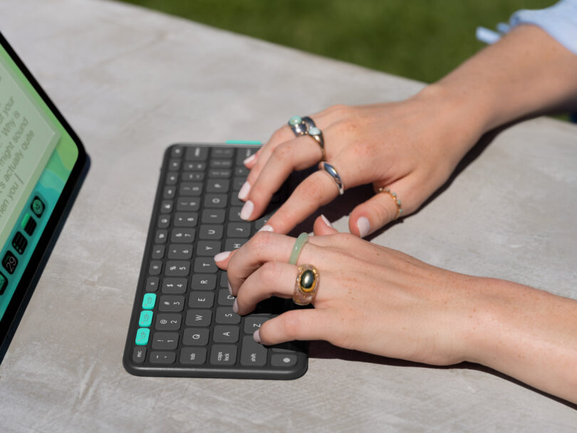 A person with rings on their fingers types on a compact wireless keyboard next to a tablet in a Flip Folio on a gray table outdoors.