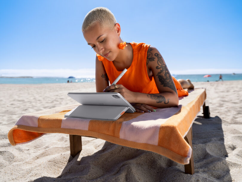 A person with short blonde hair and tattoos lies on a beach chair, using a tablet and Flip Folio with a stylus; the beach and ocean are visible in the background.