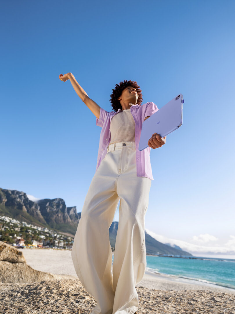A person holding a laptop with a Flip Folio stands on a rocky shore, raising one arm, with mountains, beach, and the ocean in the background under a clear blue sky.