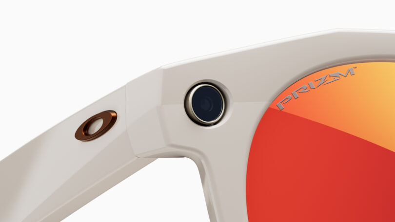 Close-up of the hinge of a white Meta sunglasses frame with an embedded small camera and orange-tinted lens labeled "Prizm.