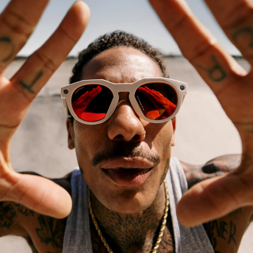 A man wearing white sunglasses with red lenses and a gray tank top looks into the camera, holding his hands up with fingers spread. His tattooed arms add an edgy vibe, creating a bold Meta-inspired aesthetic.