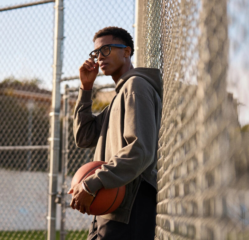 A person in glasses and a hoodie holds a basketball while leaning against a chain-link fence on an outdoor court, lost in meta reflection before their next game.