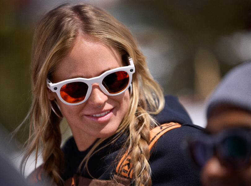 A woman with blonde braided hair, sporting Meta-inspired white-framed sunglasses and a dark hoodie, looks to the side while outdoors.