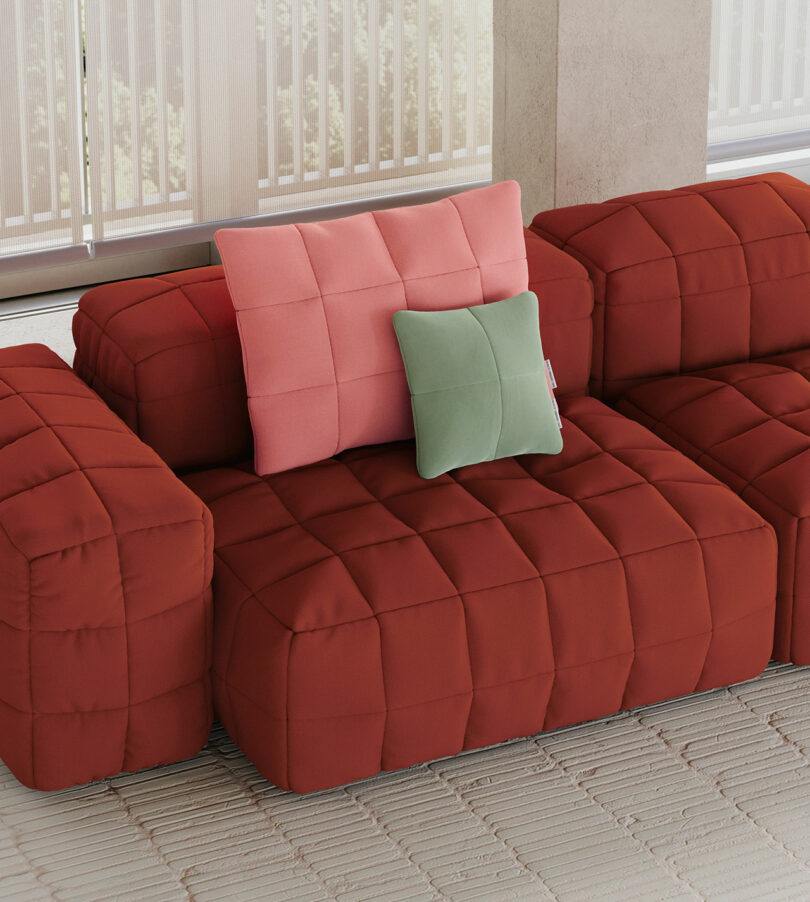 A red, quilted sectional sofa by Objekte Unserer Tage with a pink pillow and a green pillow on top, placed on a beige textured rug near large windows with vertical blinds.