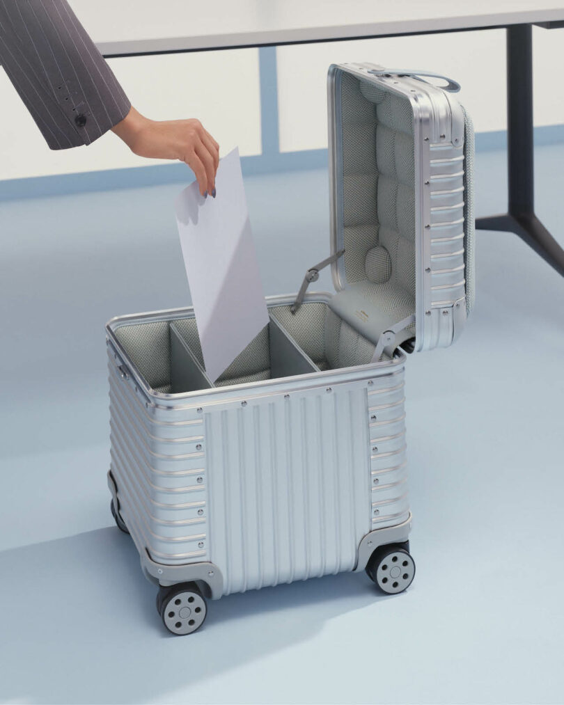 A person places a sheet of paper into an open silver suitcase with interior dividers on the floor next to a table