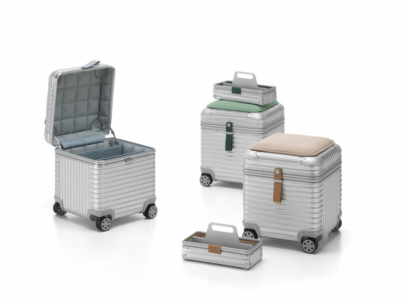 Four silver, square-shaped Vitra Rimowa suitcases with wheels and straps; one is open showing the interior, one has a tray on top, and two feature cushioned lids in green and beige.
