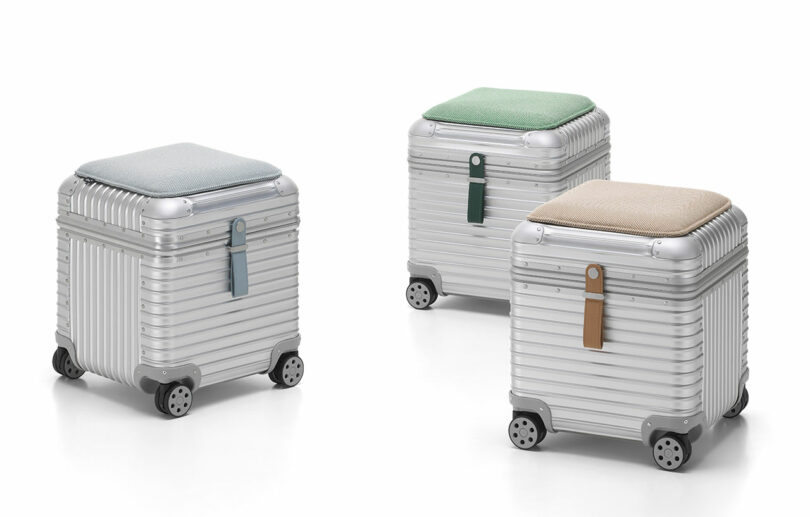 Three metallic, wheeled storage boxes from the Vitra Rimowa collection feature ribbed surfaces and cushioned tops in blue, green, and beige, each finished with a leather strap on the front.