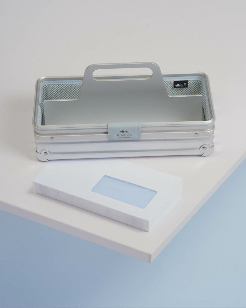 A silver metallic storage basket with a handle and mesh sides sits next to a white rectangular box with a clear window on a white and blue surface