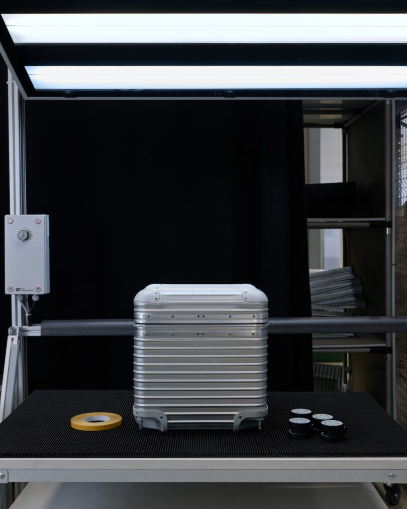 A silver hard-shell suitcase is positioned under bright lights on a dark workstation, with tape and small containers placed nearby