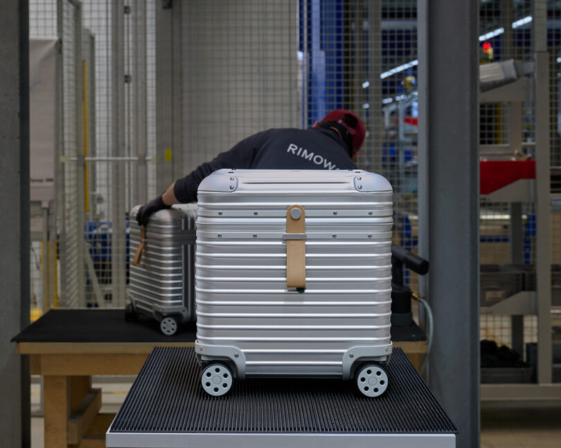 A person in a factory setting assembles or inspects silver hard-shell suitcases with wheels and leather handles on a workstation