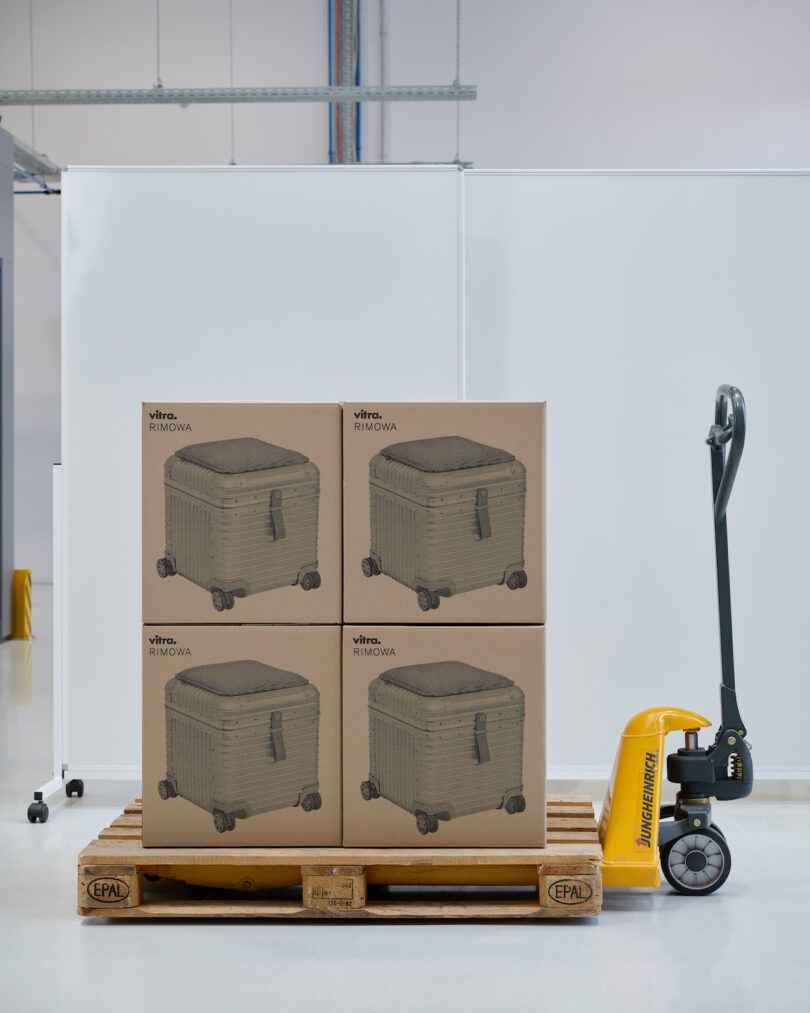 A pallet jack holds a wooden pallet stacked with four cardboard boxes featuring suitcase illustrations, situated in a clean, industrial setting