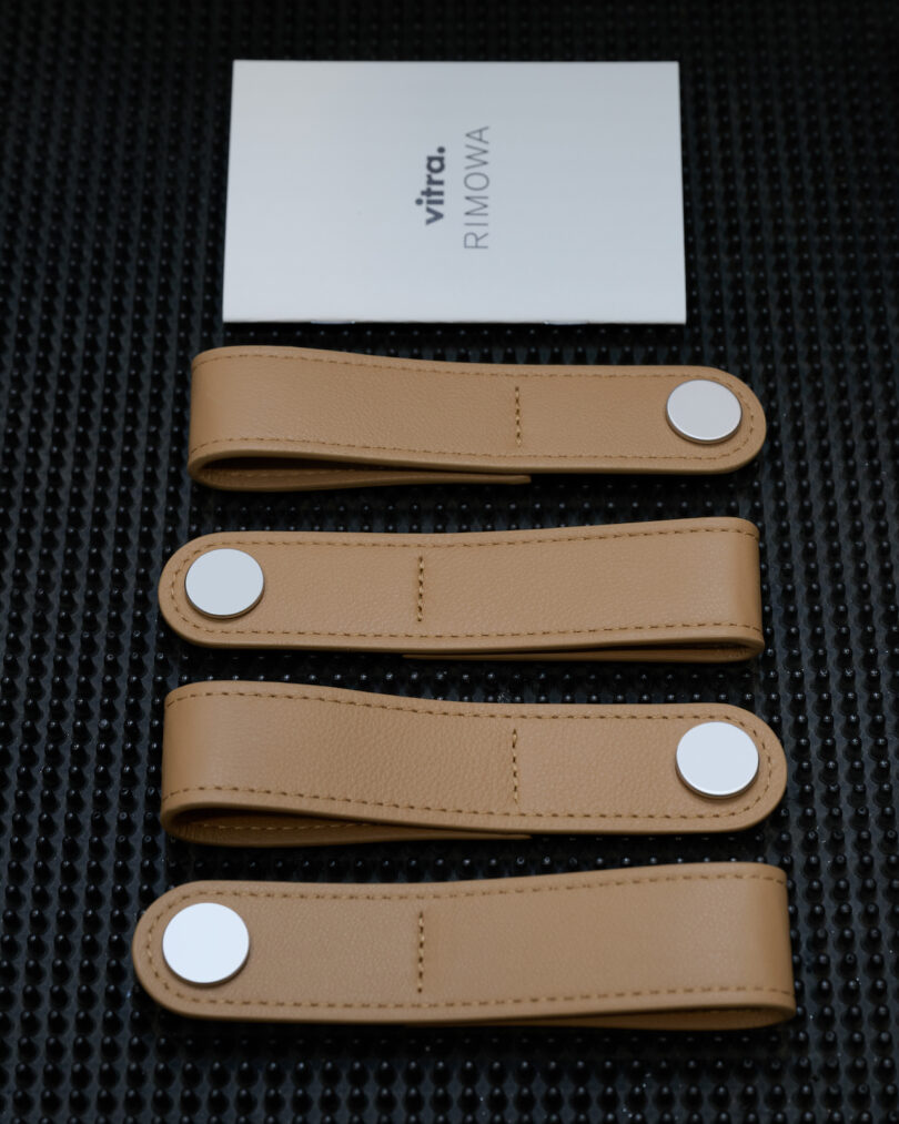 Four tan leather straps with silver snaps are laid out in a row below a white card labeled "vitra. RIMOWA" on a black textured surface