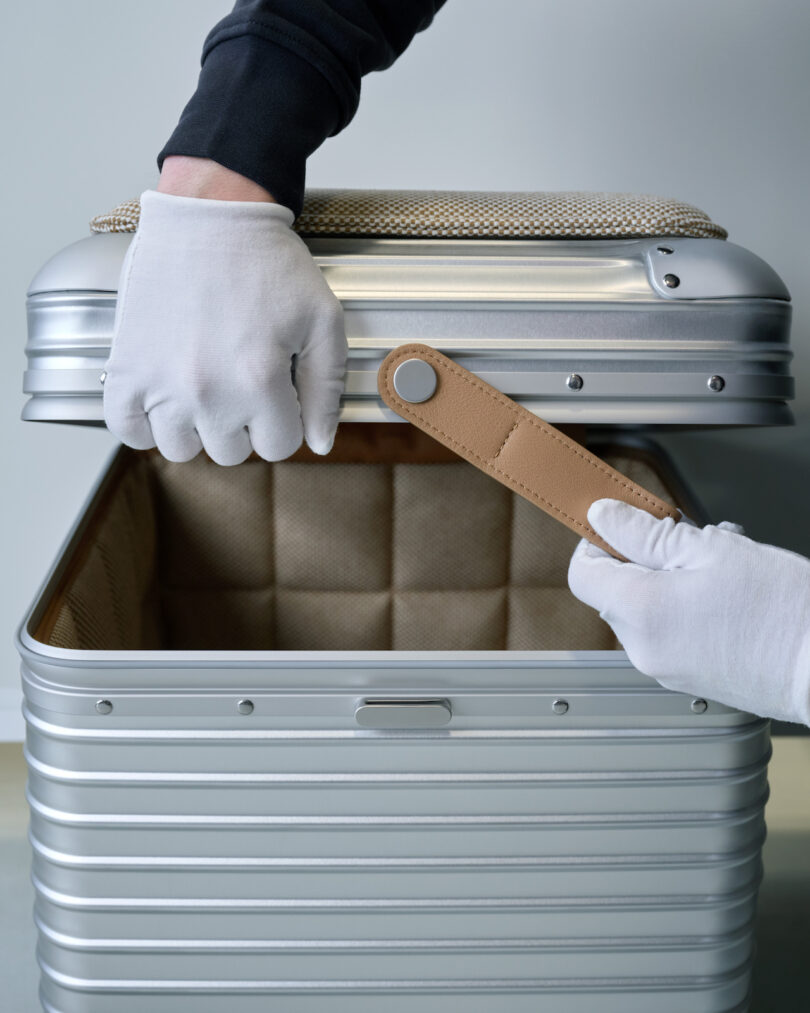 Two gloved hands are opening a silver suitcase, one lifting the lid and the other holding a tan leather strap inside the suitcase